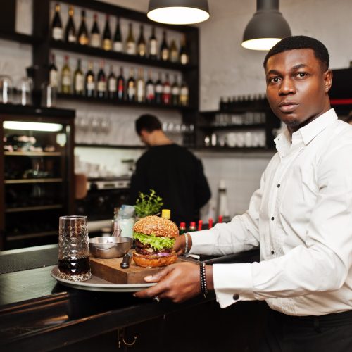 un jeune garcon afro americain tient un plateau avec un hamburger au bar du restaurant min
